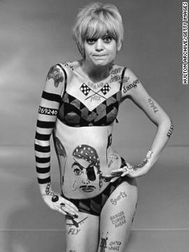 Απεικονίζεται μία ξανθιά γυναίκα, φοράει ένα μπικίνι μαγιό με γεωμετρικά σχήματα και το σώμα της έχει τατουάζ