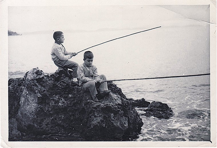Απεικονίζεται ο Γιώργος Καμίνις με ένα φίλο του καθισμένοι στα βράχια και ψαρεύουν