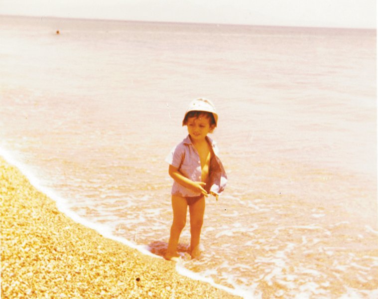 Απεικονίζεται ο Μάνος Βουλαρίνος με ένα καπελάκι και ανοιχτό πουκαμισάκι όταν ήταν παιδί σε παραλία της Άνδρου
